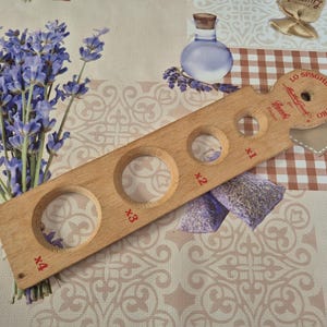 May include: A wooden spaghetti measure with four circular holes of varying sizes, labelled x1 to x4. The measure is stamped with text and a logo. The image also includes lavender sprigs, a small glass bottle, and a gingham pattern.
