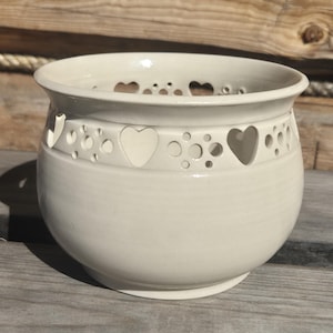 Könnte beinhalten: Ein cremefarbener Keramik-Blumentopf mit rundem Körper und breitem Rand. Der Topf hat ein dekoratives Band aus Herz- und Kreisausschnitten in der Mitte. Er steht auf einer Holzoberfläche.