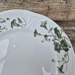 May include: White ceramic serving dish with a scalloped edge and a floral vine design in shades of green. The design features flowers and leaves. The dish is set against a rustic wooden background.