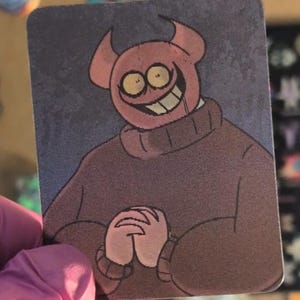 May include: A rectangular sticker featuring a cartoon illustration of a pink, horned character with wide yellow eyes and a broad, toothy grin. The character wears a brown turtleneck sweater and has hands clasped together against a dark gray background.