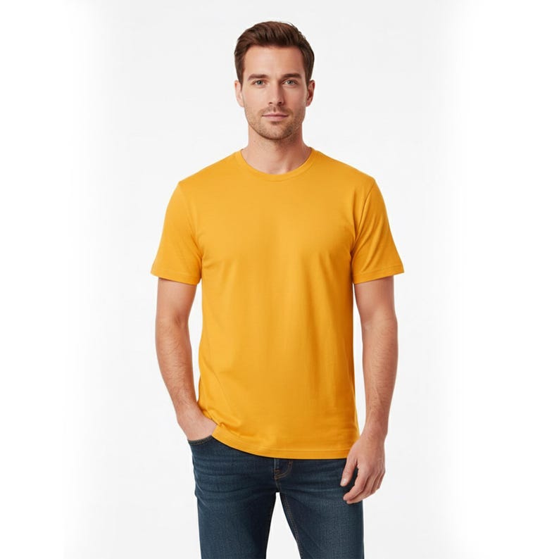 May include: A mustard yellow crew neck t-shirt. The short-sleeved shirt is made of a soft material. The person in the image is wearing blue jeans. The background is white.