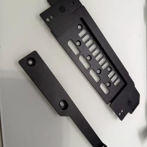 以下が含まれることがあります： 白い表面に、2つの黒い金属製のコンピューター部品が表示されています。1つは、複数の切り欠きスロットと穴がある長い長方形の部品です。もう1つは、単一の穴がある、より小さなL字型の部品です。