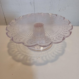 May include: A clear, light pink glass cake stand with a scalloped edge. The round plate has a textured design with a central column for support. The stand casts a shadow on the white surface.