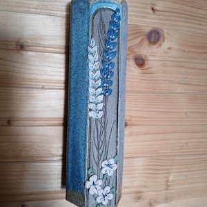 May include: A tall, hexagonal ceramic wall vase with a blue glazed finish. The center panel features a textured gray surface decorated with raised, sculpted wheat stalks in white and blue, and three small white flowers with green leaves at the base.