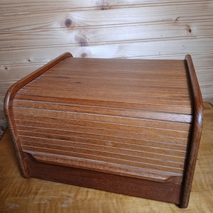 May include: A rectangular wooden bread box featuring a roll-top tambour door with horizontal slats. The container is constructed from medium-toned wood with a visible grain pattern, showcasing a curved frame design on both side panels.