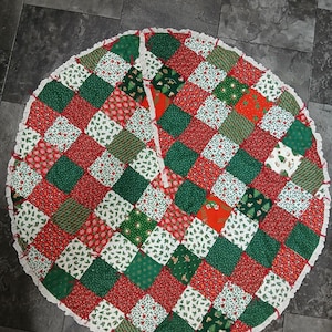 May include: A circular Christmas tree skirt featuring a patchwork design of red, green, and white fabric squares with various holiday-themed patterns. The edge is finished with a white crocheted trim, and a slit runs from the center to the edge.
