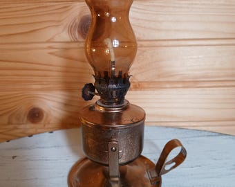 Puede incluir: Una lámpara de aceite de estilo vintage con base de metal en tono cobre, bandeja circular y una pequeña asa lateral. La lámpara incluye una chimenea de cristal de color ámbar oscuro y un mecanismo de quemador central con una mecha blanca.