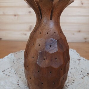 May include: A handcrafted wooden pineapple-shaped decorative item. The brown wood has a faceted design with small holes and a crown of leaf-like elements. It sits on a white lace doily, with a wooden background.