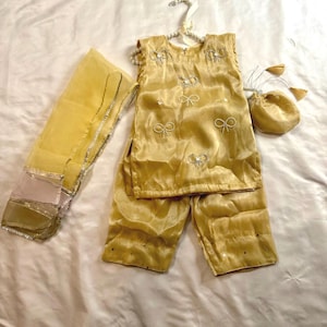 May include: A child's outfit in a golden yellow hue, comprising a sleeveless top, trousers, a scarf, and a small purse. The top and trousers feature a quilted design and bow-shaped decorations. The scarf is trimmed with silver.