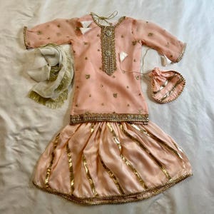 May include: A peach-colored traditional outfit with gold embellishments. The set includes a top, a skirt, a scarf, and a small pouch. The top and skirt are adorned with gold trim and accents. The scarf has gold fringe.