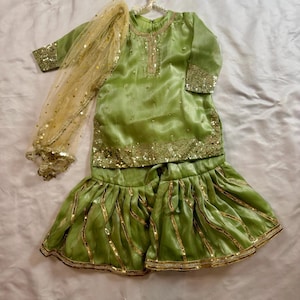 May include: A lime green, embellished, two-piece outfit with a matching sheer scarf. The top has a round neckline and three-quarter sleeves. The flared pants are adorned with gold stripes. The scarf is a sheer, gold-toned fabric with gold sequins.
