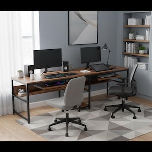 May include: A long, rectangular double workstation desk with a dark wood-grain surface and a black metal frame. The desk features two computer monitors, two sets of speakers, a keyboard, a mouse, and a drawing tablet. Two office chairs are present.