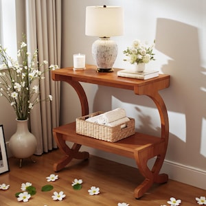 May include: A two-tiered wooden console table with a warm, reddish-brown finish. The top shelf holds a lamp, candle, and flowers. The lower shelf features a woven basket with rolled towels. The table has curved supports and is set on a hardwood floor.