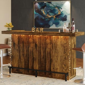 May include: A rustic wooden bar unit featuring a dark brown wood grain finish, a black metal footrest, and warm string lights draped along the front edge. The top surface holds a gold-colored light-up sign spelling BAR and a glass bottle set.