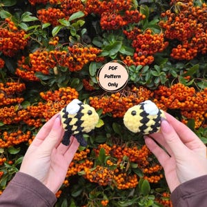 May include: Two crocheted bee toys with yellow and black checkered bodies and white wings are held in front of a backdrop of orange berries and green leaves. The text "PDF Pattern Only" is in a circle.