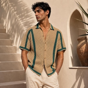 May include: A tan crochet shirt with dark green trim and a button-down front. The short-sleeved shirt is paired with light-colored pants. The image is taken outdoors with a neutral background.