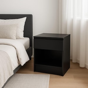 May include: A black nightstand with a drawer and an open shelf. The nightstand is rectangular and sits on a light-colored floor. The bed frame and bedding are visible in the background, with a neutral-colored rug in front of the bed.