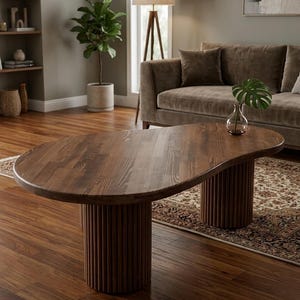 May include: A dark brown, oval-shaped wooden coffee table with two cylindrical, ribbed legs. The table sits on a hardwood floor in a living room setting. A glass vase with a green leaf sits on the table.