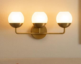 Puede incluir: Aplique de pared de tres luces con acabado en latón. La lámpara tiene tres pantallas esféricas de vidrio esmerilado. El aplique está montado sobre una cama con cojines naranjas y blancos. El diseño es moderno y elegante.