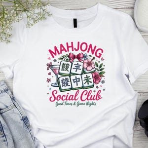 May include: White t-shirt featuring a colorful graphic design. The design includes the word "MAHJONG" in pink, Mahjong tiles, flowers, and the words "Social Club" and "Good Times & Game Nights".