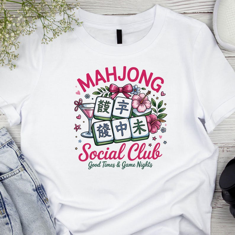 May include: White t-shirt featuring a colorful graphic design. The design includes the word "MAHJONG" in pink, Mahjong tiles, flowers, and the words "Social Club" and "Good Times & Game Nights".