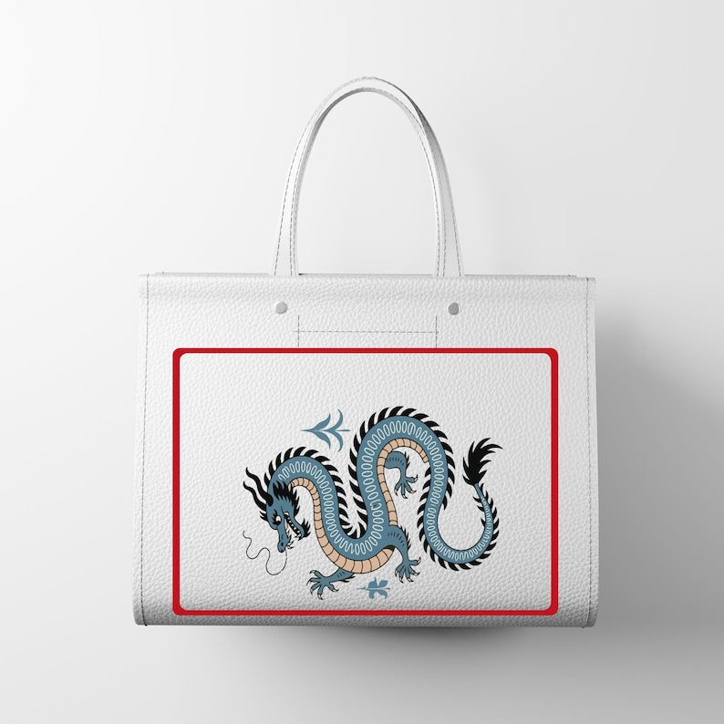 Puede incluir: Bolso tote blanco con un dise&ntilde;o de drag&oacute;n azul y negro dentro de un borde rojo. El drag&oacute;n tiene un cuerpo largo y sinuoso. El bolso tiene dos asas blancas. El bolso es de material texturizado.