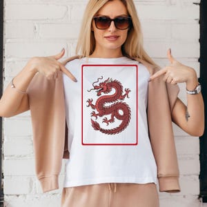 Puede incluir: Camiseta blanca con un dise&ntilde;o de drag&oacute;n rojo dentro de un marco rectangular rojo. El drag&oacute;n est&aacute; detallado con un cuerpo curvado y l&iacute;neas fluidas. La camiseta es de estilo cuello redondo cl&aacute;sico.