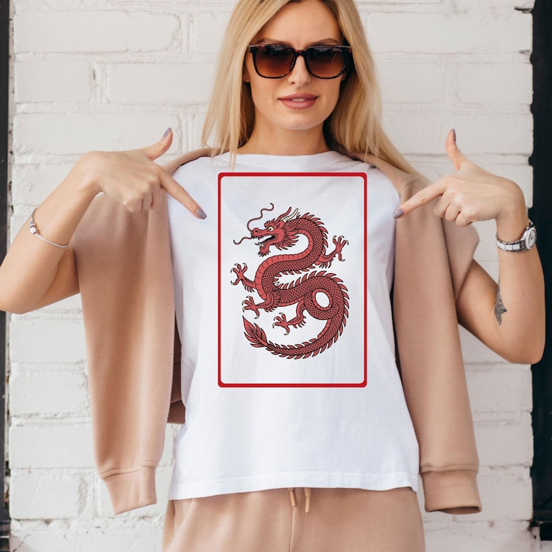 Puede incluir: Camiseta blanca con un dise&ntilde;o de drag&oacute;n rojo dentro de un marco rectangular rojo. El drag&oacute;n est&aacute; detallado con un cuerpo curvado y l&iacute;neas fluidas. La camiseta es de estilo cuello redondo cl&aacute;sico.