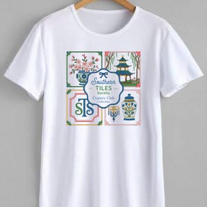 May include: White t-shirt featuring a colorful graphic design. The design includes floral elements, a pagoda, and the text "Southern Tiles Society Country Club Collection". The shirt has a classic crew neck and short sleeves.