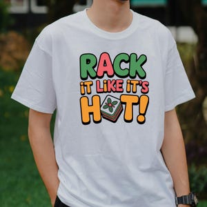 May include: White t-shirt with the text "RACK IT LIKE IT'S HOT!" in green, pink, and orange. The design includes a graphic of a stack of food with a flower on top. The shirt is a casual style.