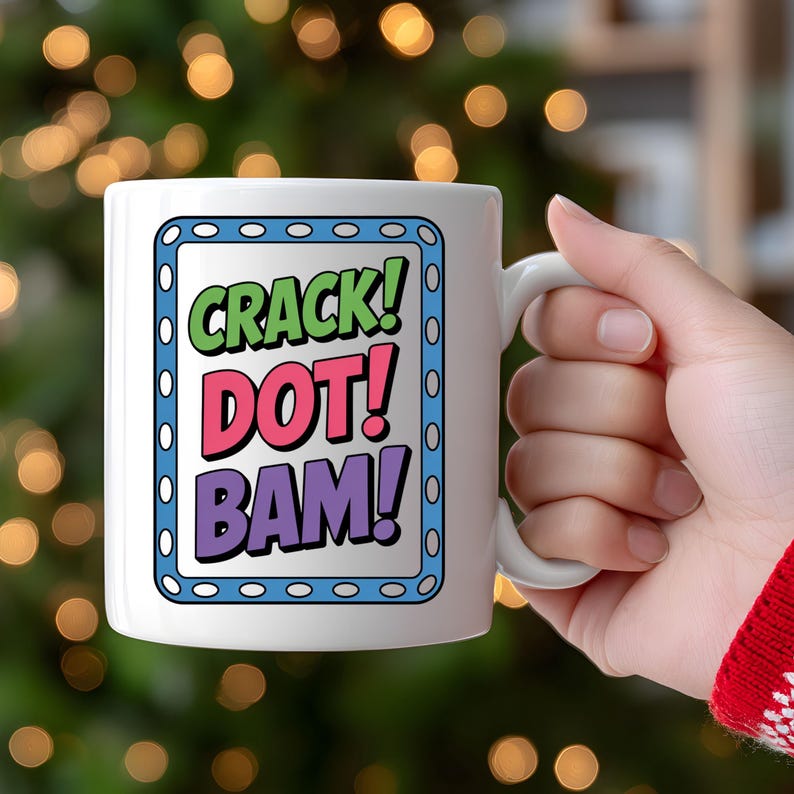 May include: White ceramic mug with the words "CRACK! DOT! BAM!" in green, pink, and purple, framed by a blue border. The mug has a white handle and is held by a hand.
