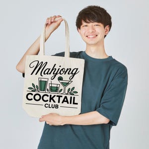 May include: Cream-colored tote bag with the words "Mahjong Cocktail Club" in black script and block letters. The design includes illustrations of cocktail glasses and foliage in green. The bag is held by a person.