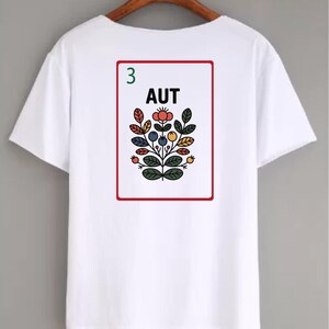 Puede incluir: Camiseta blanca con un dise&ntilde;o floral colorido dentro de un marco rectangular rojo. El dise&ntilde;o incluye la palabra "AUT" y el n&uacute;mero "3". Los elementos florales son en tonos de rojo, azul, amarillo y verde.