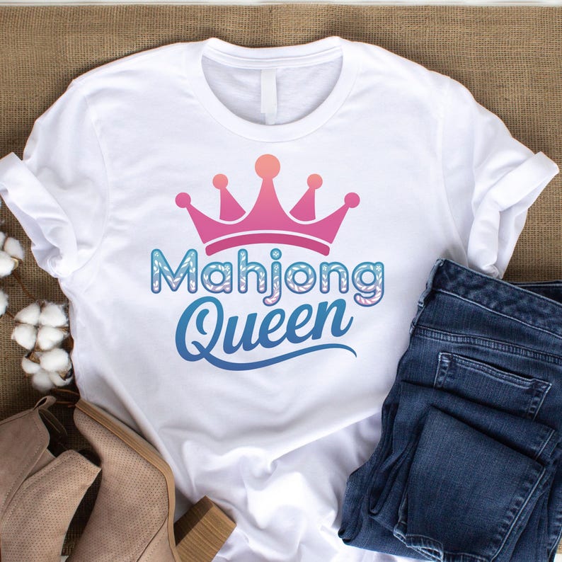 May include: White t-shirt with a pink and orange crown design above the words "Mahjong Queen" in blue and white. The shirt is paired with blue jeans and tan boots, suggesting a casual outfit.