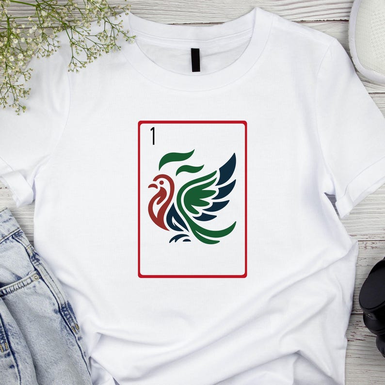 Puede incluir: Camiseta blanca con un dise&ntilde;o de carta de juego que presenta un p&aacute;jaro estilizado en rojo, verde y azul. La carta tiene un borde rojo y el n&uacute;mero "1". El dise&ntilde;o es un gr&aacute;fico vibrante y llamativo.