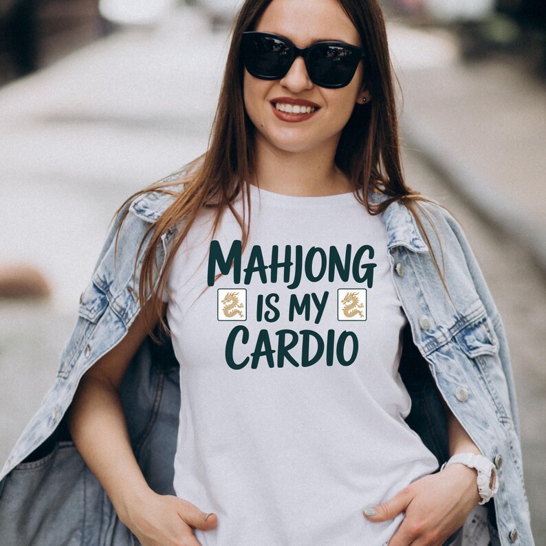 May include: White t-shirt with the text "MAHJONG IS MY CARDIO" in dark green. The shirt features two golden dragon symbols. The person in the image is wearing sunglasses and a denim jacket.