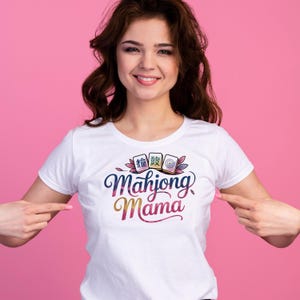 May include: White t-shirt with a colorful "Mahjong Mama" graphic. The design includes stylized Mahjong tiles and floral accents. The text is in a script font with a gradient of colors.