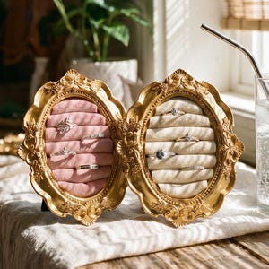 Könnte beinhalten: Eine goldfarbene Schmuckschatulle, geöffnet, um Ringe auf rosa Samt zu präsentieren. Die Schatulle steht auf einer Holzoberfläche neben einem Glas Wasser und einer Parfümflasche. Der Hintergrund zeigt ein Fenster mit einem Bambusrollo.