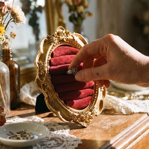 Könnte beinhalten: Eine goldgerahmte Schmuckauslage mit rotem Samtinterieur hält einen silbernen Ring. Die Auslage steht auf einer Holzkommode mit einem Spitzentischläufer. Getrocknete Blumen in Glasflaschen und eine kleine Schale mit Ringen sind ebenfalls zu sehen.