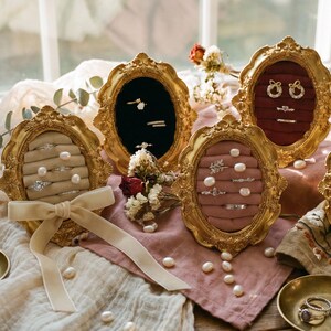 Könnte beinhalten: Eine Auswahl goldgerahmter Ringdisplays mit Samtinterieur in verschiedenen Farben, die Ringe mit Perlen und Edelsteinen präsentieren. Ein cremefarbenes Band und kleine Messingschalen mit Ringen sind ebenfalls vorhanden, was eine elegante Schmuckpräsentation ergibt.
