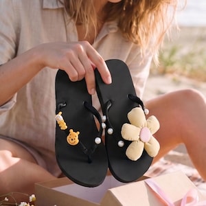 Op de afbeelding: Zwarte slippers met decoratieve versieringen. Eén heeft een gele bloem met een roze centrum en witte parels, de andere een gele beer-bedel. De slippers worden vastgehouden door een persoon die op het strand zit.