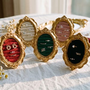 Könnte beinhalten: Fünf verzierte, goldgerahmte Ringdisplays mit ovalen Öffnungen. Jeder Rahmen enthält Ringe und Ohrringe auf Samt in verschiedenen Farben: Rot, Grün, Rosa, Creme und Schwarz. Die Displays sind auf einer weißen Oberfläche angeordnet.