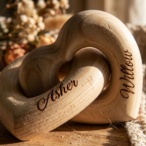 May include: A wooden heart-shaped toy with the names "Asher" and "Willow" engraved on it. The two hearts are interlocked, and the wood has a natural, light color. The background includes dried flowers and a textured surface.
