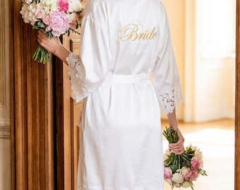 May include: A white satin bride's robe with lace trim and the word "Bride" embroidered in gold script on the back. The robe is worn by a person holding two bouquets of pink and white flowers.