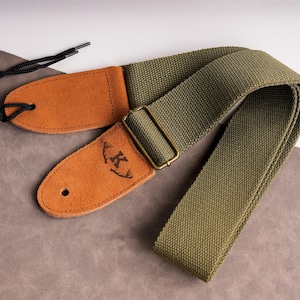 May include: Olive green guitar strap with brown leather ends. The leather ends have a debossed design with the letter "K" and a deer antler motif. The strap has a metal buckle and black cord.