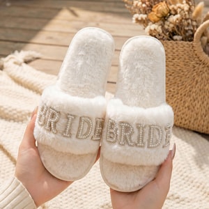 May include: A pair of white, fluffy slippers with the word "BRIDE" spelled out in silver-colored letters and small pearls. The slippers are held in a person's hands, with a woven handbag and dried flowers in the background.