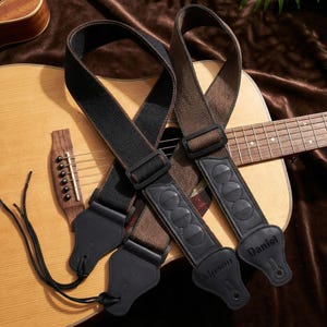 May include: Two guitar straps, one black and one brown, rest on a light brown acoustic guitar. The straps have black leather ends and are personalized with the names "Mason" and "Daniel". The guitar has a wooden bridge and strings.