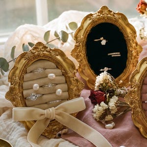 Könnte beinhalten: Eine Auswahl goldgerahmter Ringdisplays mit Samtinterieur in verschiedenen Farben, die Ringe mit Perlen und Edelsteinen präsentieren. Ein cremefarbenes Band und kleine Messingschalen mit Ringen sind ebenfalls vorhanden, wodurch eine elegante Schmuckpräsentation entsteht.