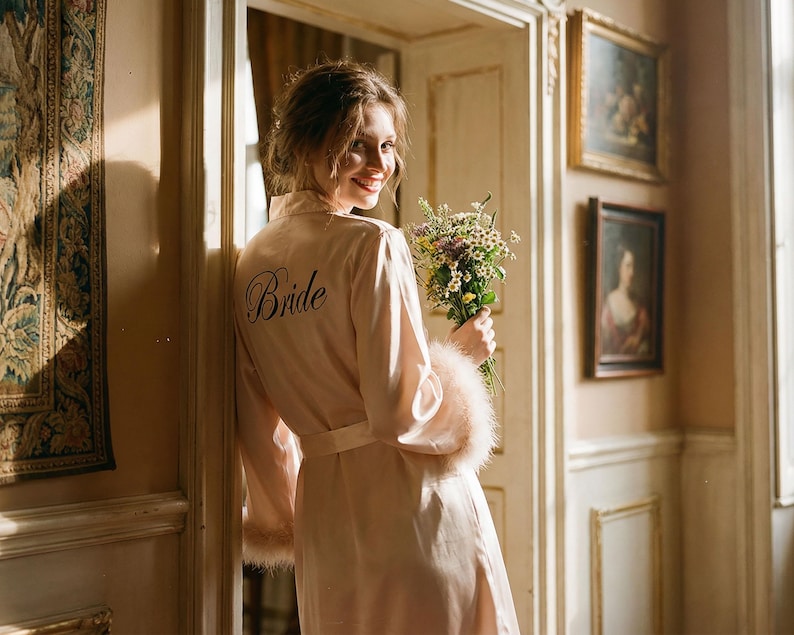May include: A pale pink satin robe with the word "Bride" embroidered on the back. The robe features fluffy feather trim on the sleeves and a matching belt. The bride is holding a bouquet of wildflowers.