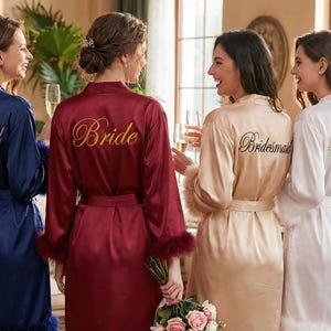 May include: Four women in satin robes of navy, burgundy, champagne, and white. The burgundy robe has "Bride" embroidered in gold, the others say "Bridesmaid". The robes have feather trim. The women are holding glasses of champagne.